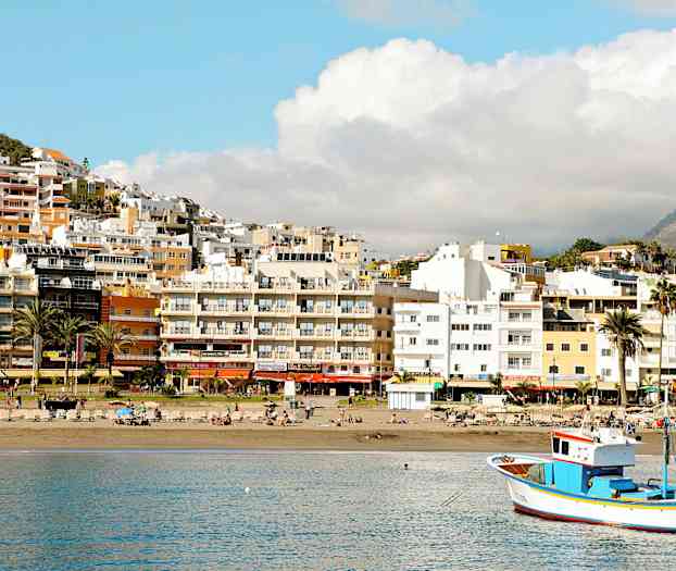 En strand med solsenger, en fiskebåt på vannet og bygninger bak. (AI-generert tekst)