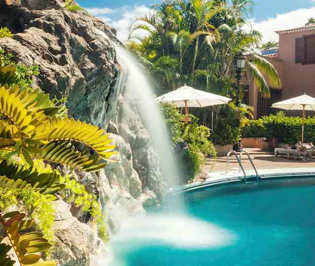 Vannfall som renner ned i et svømmebasseng, omgitt av grøntområder og solsenger. (AI-generert tekst)