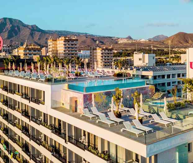 Hotellbygning med et infinitybasseng og solsenger på takterrassen, i bakgrunnen fjell og byer. (AI-generert tekst)