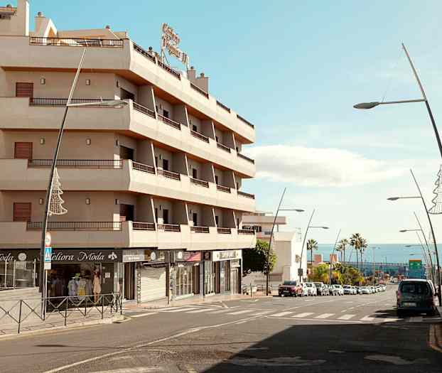 En bygning med balkonger ved en gate med biler og gatelys, med havet i bakgrunnen. (AI-generert tekst)