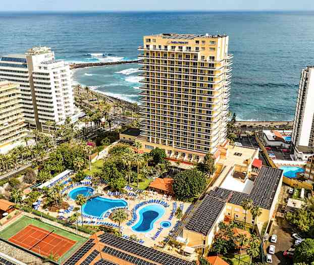 Hotellanlegg med bassengområde og tennisbane, ved sjøen, omgitt av høye bygninger. (AI-generert tekst)