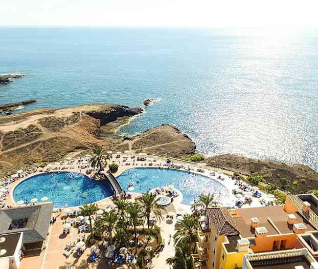 Flyfoto av en resort med basseng, solsenger og havutsikt. (AI-generert tekst)
