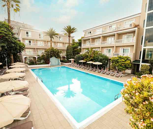 Hotellområde med et utendørs basseng omgitt av solsenger og parasoller samt bygninger med balkonger. (AI-generert tekst)