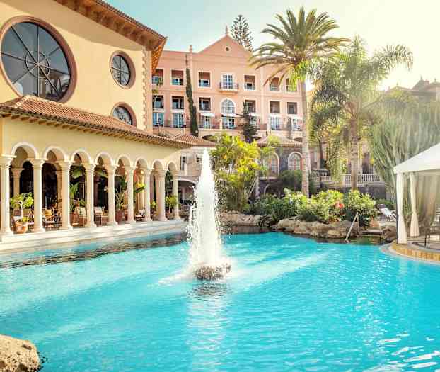 Hotellbygning med basseng og fontene i forgrunnen, omgitt av palmer og grøntområder. (AI-generert tekst)