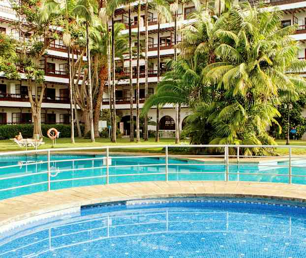 Utendørsbasseng foran en hotellbygning med balkonger og frodige trær. (AI-generert tekst)