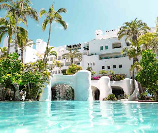 Hvit bygning med palmer rundt og basseng i forgrunnen. (AI-generert tekst)