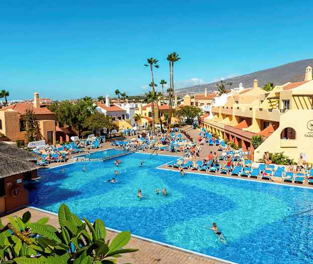 Stor utendørsbasseng omgitt av solsenger og bygninger med fjell i bakgrunnen. (AI-generert tekst)