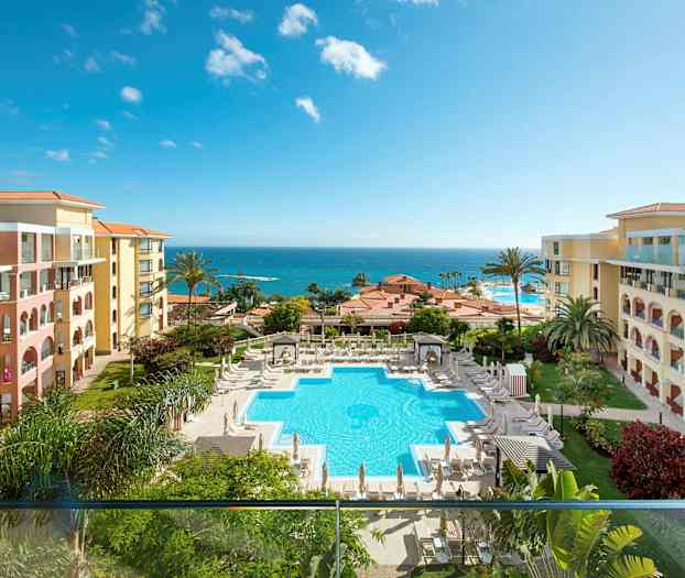 Bilde av et hotellområde med et basseng, omgitt av solsenger og parasoller. Hotellbygningene ligger på begge sider, og i bakgrunnen vises havet. (AI-generert tekst)