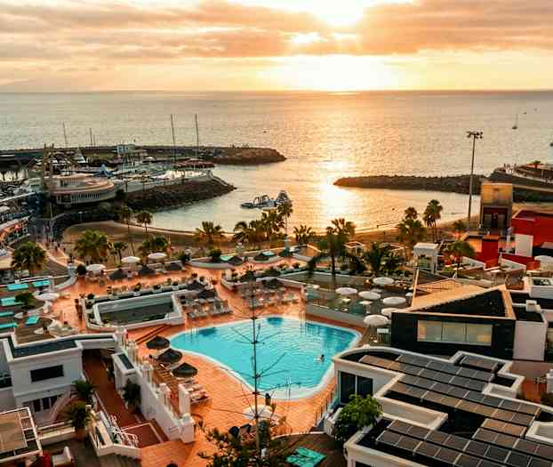 Utsikt over et kystområde ved solnedgang med et anlegg som har basseng og solterrasse i forgrunnen samt havet og småbåter i bakgrunnen. (AI-generert tekst)