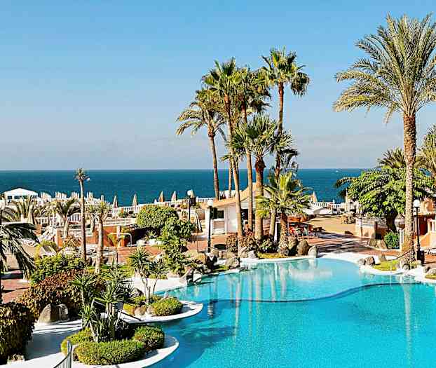 En ferieresort med et basseng, omgitt av palmer og med utsikt over havet i bakgrunnen. (AI-generert tekst)