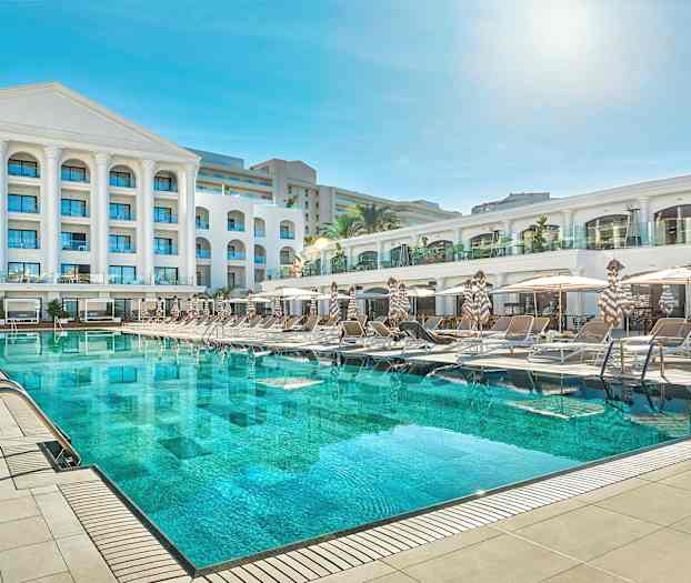 En hotellbygning med et stort svømmebasseng og solsenger foran. (AI-generert tekst)