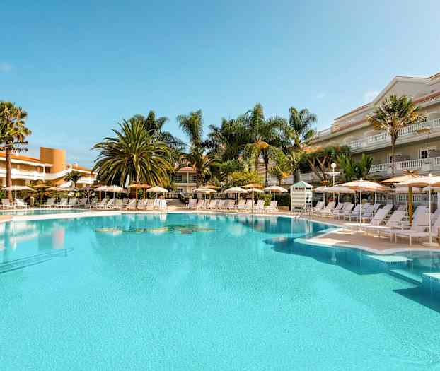 Utendørs basseng med solsenger og parasoller omgitt av palmer og bygninger. (AI-generert tekst)