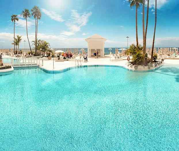 Et stort utendørs basseng omgitt av solsenger og palmer med havet i bakgrunnen. (AI-generert tekst)