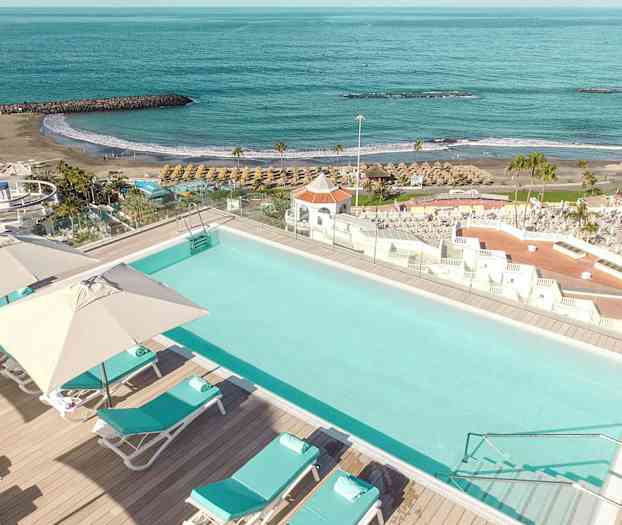 Et basseng med solsenger og parasoller, som ligger nær en strand og havet. (AI-generert tekst)