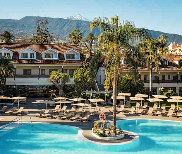 Et feriested med bassengområde omgitt av solsenger og parasoller. Tilstøtende bygninger med røde tak og fjell i bakgrunnen. (AI-generert tekst)