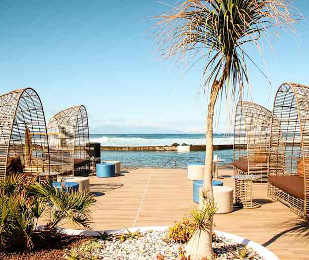 Utemøbler med rottingdesign står på en treplattform ved havet. (AI-generert tekst)