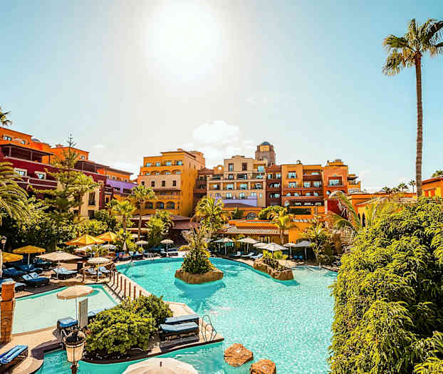 Ferieanlegg med basseng, solsenger og fargerike hotellbygg under blå himmel. (AI-generert tekst)