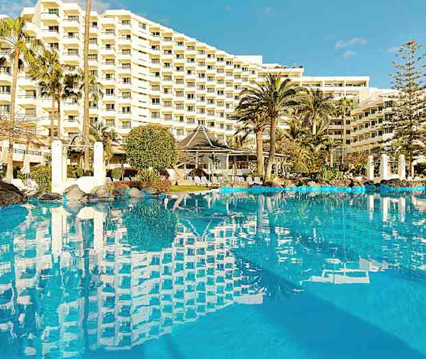 Et bilde av et utendørs svømmebasseng omgitt av palmer, med et stort hotell i bakgrunnen. (AI-generert tekst)