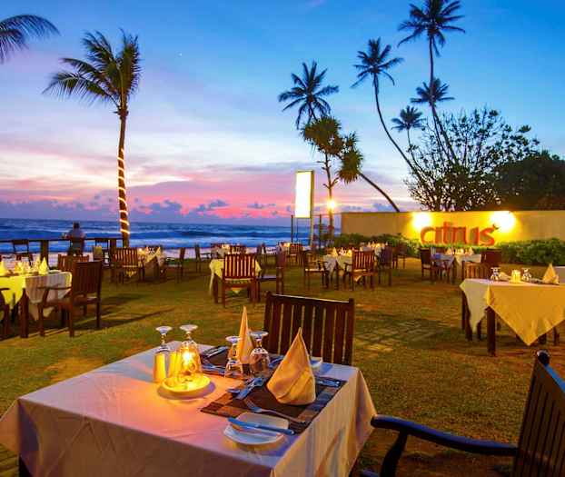 En utendørs restaurant med hvite duker og trestoler ved solnedgang nær sjøen. (AI-generert tekst)