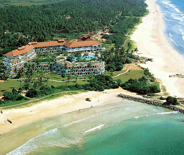 Luftfoto av et hotellkompleks omgitt av grøntområder ved kysten med en lang sandstrand og hav. (AI-generert tekst)