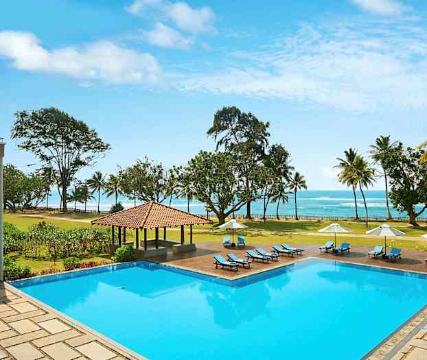 Et utendørs svømmebasseng med solsenger og parasoller, omgitt av en plen og trær, med utsikt over stranden og havet. (AI-generert tekst)