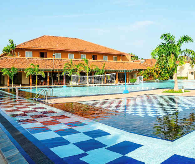 Et stort utendørsbasseng med flislagt fargerikt område, omgitt av palmer og bygninger med teglfargede tak. (AI-generert tekst)