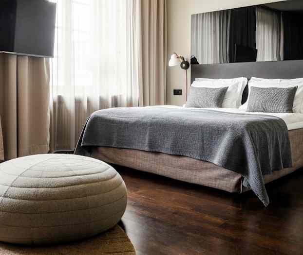 Et moderne hotellrom med en stor seng, mørkt tregulv og en grå sittepuff. (AI-generert tekst)