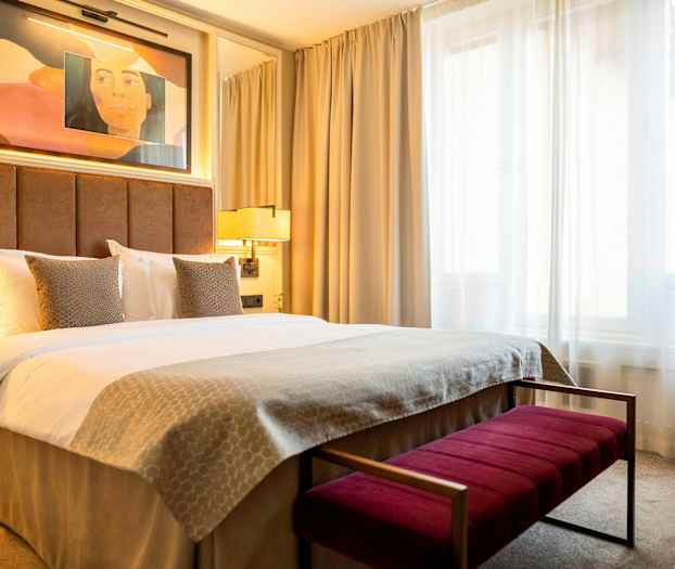 Et hotellrom med en dobbeltseng, beige sengetøy og to puter foran en polstret brun sengegavl. Lamper og et vindu med lyse gardiner vises i bakgrunnen. (AI-generert tekst)