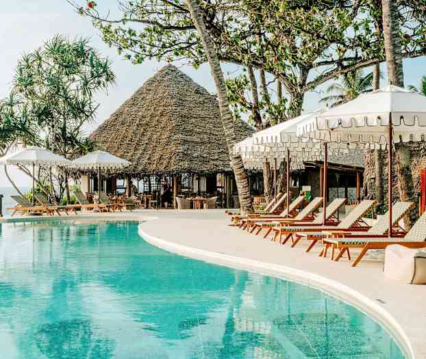 Et bassengområde med solsenger og parasoller. I bakgrunnen er en bygning med stråtak og palmer. (AI-generert tekst)