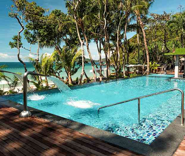 Et basseng med klart blått vann, omgitt av tropiske trær og med utsikt over havet. (AI-generert tekst)