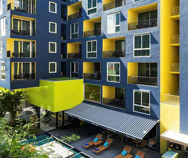 Bygning med blå og gule fasader samt balkonger, foran et basseng og solsenger. (AI-generert tekst)