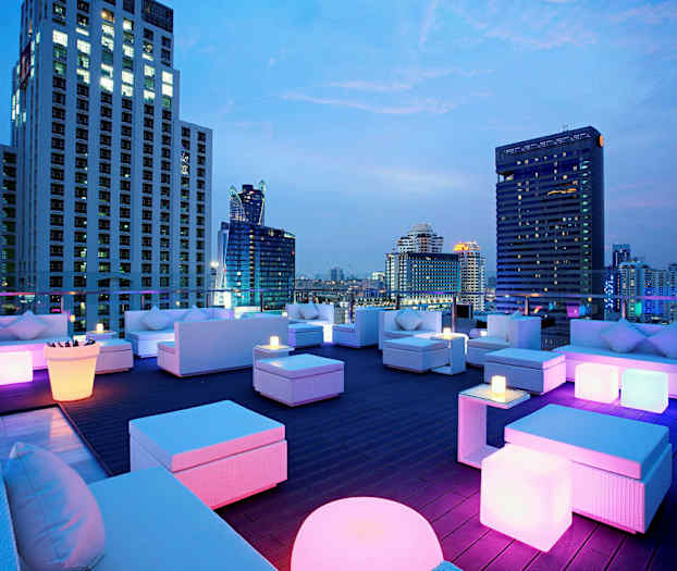 En takterrasse med moderne møbler og fargerik belysning, omgitt av høye bygninger om kvelden. (AI-generert tekst)
