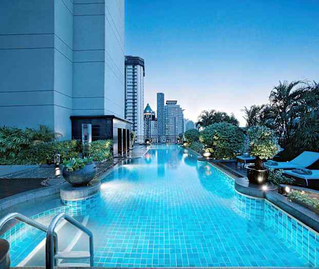 Et utendørsbasseng ved en bygning omgitt av vegetasjon, med solsenger og høye bygninger i bakgrunnen. (AI-generert tekst)