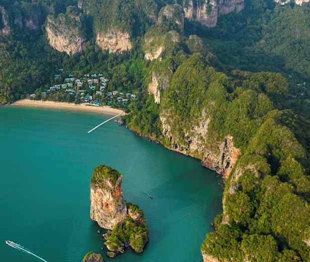 Luftfoto av en tropisk kyst med en strand, klipper og grønn vegetasjon, samt hus synlige mellom trærne. (AI-generert tekst)