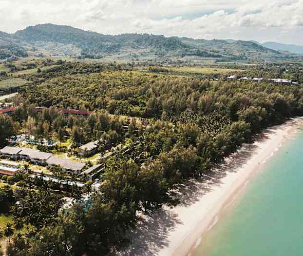 Flyfoto av en strand med frodige trær og bygninger i bakgrunnen. (AI-generert tekst)