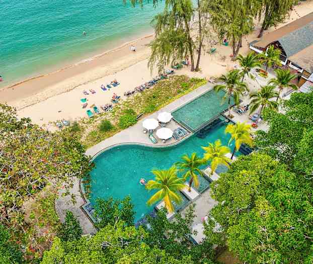 Et flyfoto av en strand med solsenger og et bassengområde omgitt av grøntområder. (AI-generert tekst)