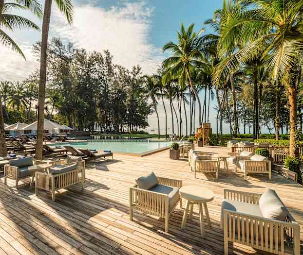 En treterrasse med grå sofagrupper og salongbord, solsenger rundt et basseng og omgivelser av palmer og trær. (AI-generert tekst)