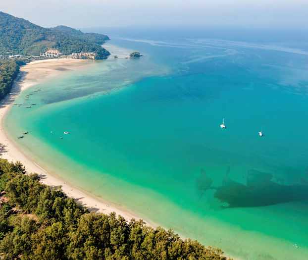 Lang hvit sandstrand mot turkisblått hav med båter og frodig omgivelser. (AI-generert tekst)