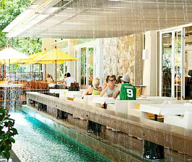 Et uteområde med bord og stoler, samt en vannkanal og gulhvite parasoller. (AI-generert tekst)