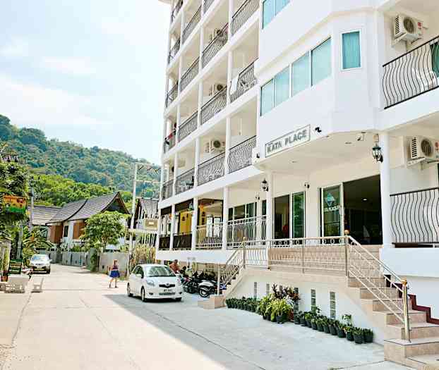 Utenfor Kata Place, en moderne bygning med balkonger, står parkerte kjøretøy og en kvinne går forbi. (AI-generert tekst)