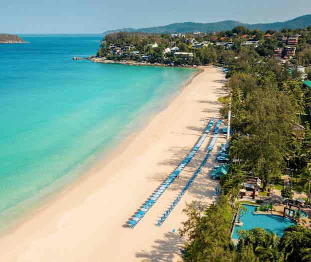 Luftfoto av en sandstrand med solsenger og parasoller langs sjøen samt grønne områder i bakgrunnen. (AI-generert tekst)