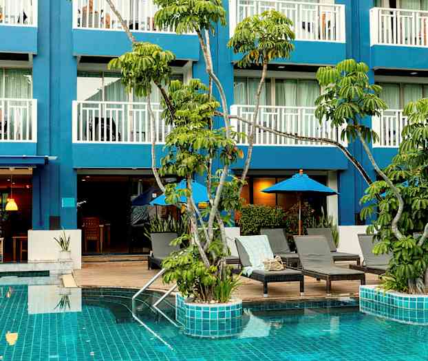 Bildet viser et basseng med turkist vann omgitt av solsenger og parasoller, samt en blå bygning med balkonger i bakgrunnen. (AI-generert tekst)