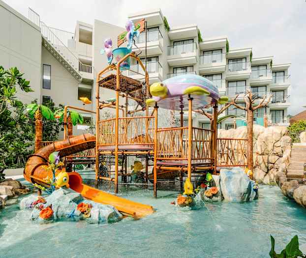 Lekeområde med vannsklie og lekeapparater, omgitt av grønne planter og steinblokker. Hotellbygning i bakgrunnen. (AI-generert tekst)