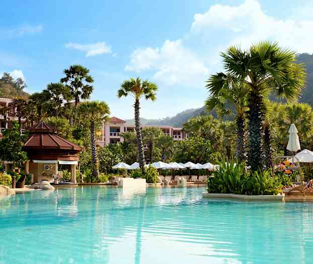 En basseng omgitt av høye palmer, solsenger med parasoller og bygninger omgitt av grøntområder. (AI-generert tekst)