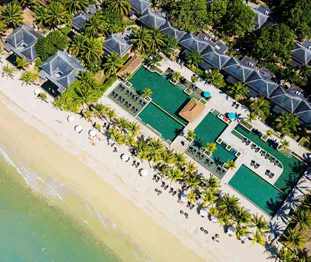 Flyfoto av en strand med parasoller, palmer, bassengområde og bygninger. (AI-generert tekst)