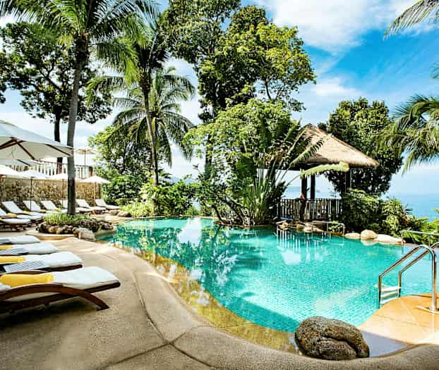 Et utendørs basseng omgitt av palmer, solsenger og parasoller, med utsikt over havet i bakgrunnen. (AI-generert tekst)