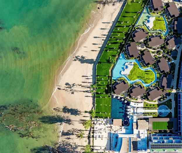 Flybilde av en strand og et hotellområde med bassenger og bygninger. (AI-generert tekst)