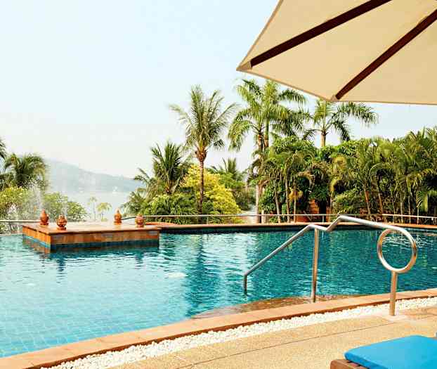 Et utendørs svømmebasseng med utsikt over grønne palmer og skogkledde åser, med solsenger og en parasoll i forgrunnen. (AI-generert tekst)