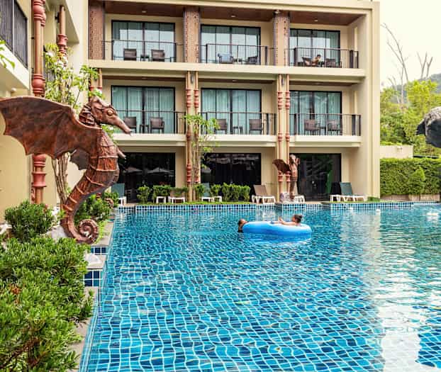 Utendørsbasseng med et barn som flyter på en blå badering, omgitt av hotellbygninger og grøntområder. (AI-generert tekst)