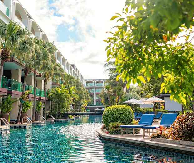 Et basseng med klart vann omgitt av solsenger, parasoller og frodige trær, nær en bygning med balkonger. (AI-generert tekst)